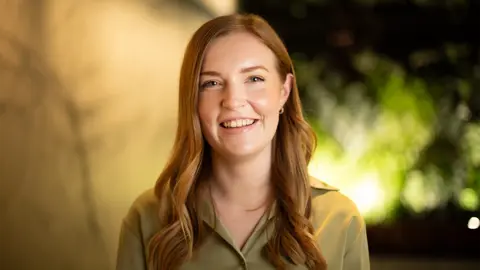 Portrait of Katie Demack, Senior Project Manager, Australia, close up wearing green shirt with greenery in background.