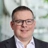 Portrait of Euan Reaper, Director, Europe, Cost Management, standing outside in front of an outdoor urban setting, featuring modern architectural elements with clean lines. There are hints of greenery, suggesting some trees or plants.