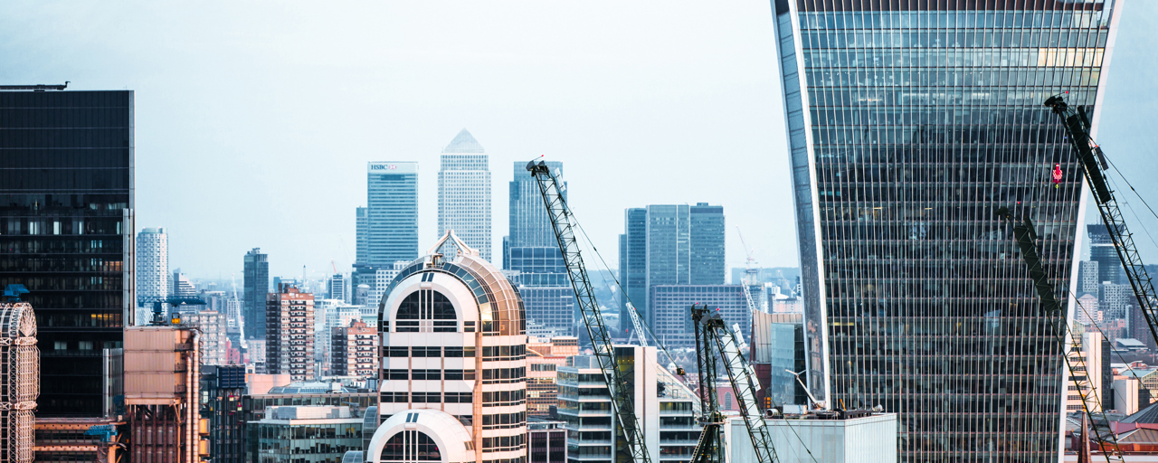 A view of the London skyline at dusk, bathed in a pinkish-golden light, with a cloudy sky. Four construction cranes are visible, hinting at ongoing construction work in the area.