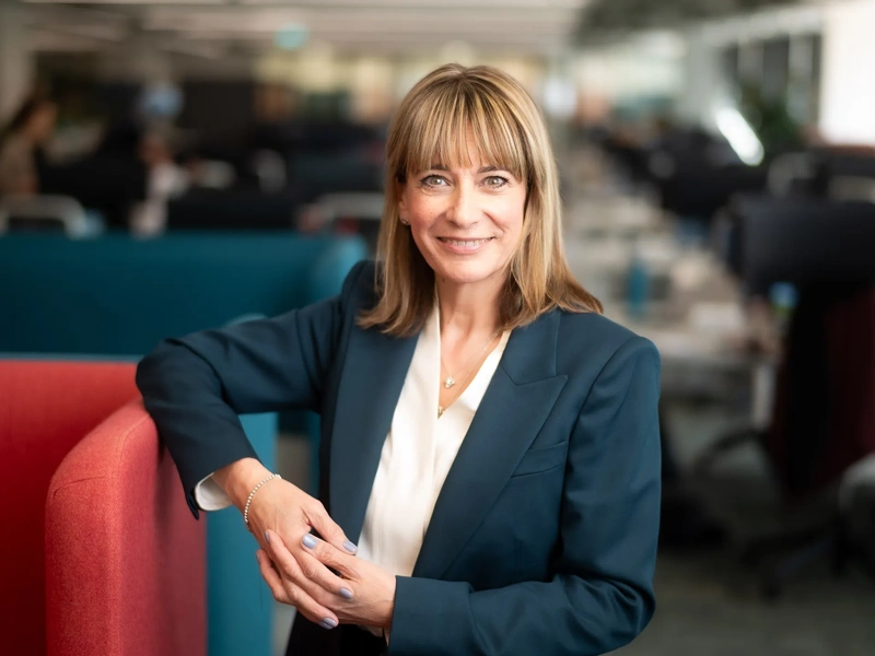 Portrait of Emma Gilthorpe, Chief Transformation Officer, standing in office against red chair in blue suit.