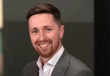 Portrait of Ruairidh Holland, Principal consultant, smiling in a grey suit against dark office background