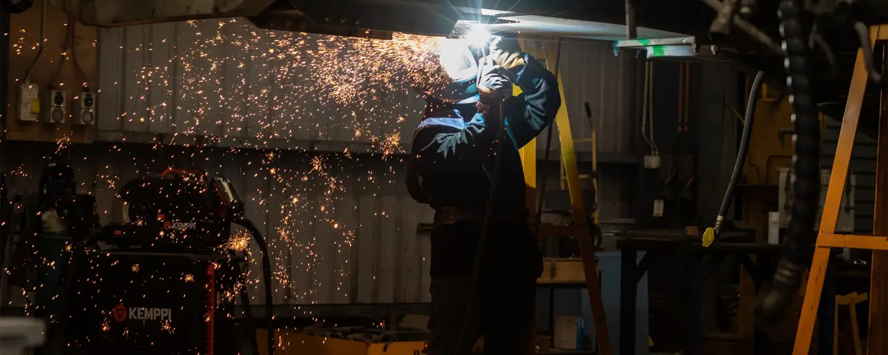 A welder in protective gear, including a welding helmet and gloves, is working in a dimly lit workshop. Bright orange sparks fly outward as they weld metal overhead, illuminating the area around them. Industrial equipment and tools are visible in the background, creating a rugged and workshop-like atmosphere