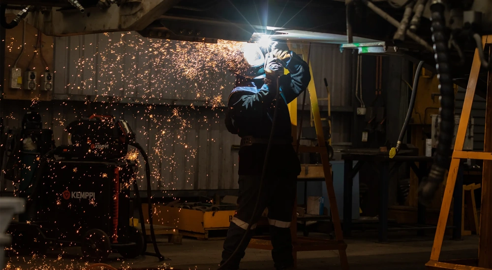 A welder in protective gear, including a welding helmet and gloves, is working in a dimly lit workshop. Bright orange sparks fly outward as they weld metal overhead, illuminating the area around them. Industrial equipment and tools are visible in the background, creating a rugged and workshop-like atmosphere