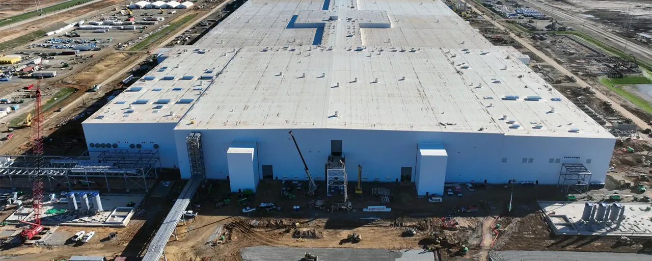 An aerial shot of a large, grey EV battery manufacturing facility surrounded by a building works. 