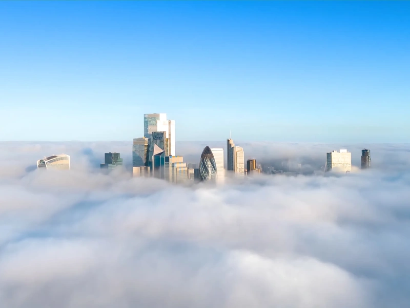 City skyline with modern skyscrappers rising above a layer of fog under a clear blue sky.