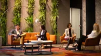 Group of four colleagues sitting separately in pairs on orange sofas in a plant-filled office.