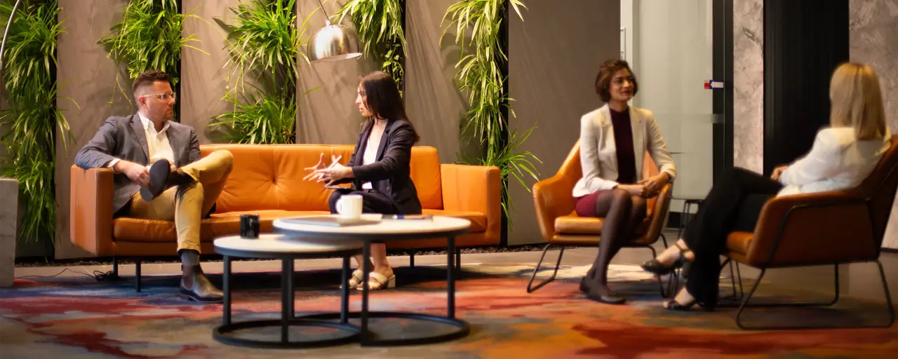 Group of four colleagues sitting separately in pairs on orange sofas in a plant-filled office.