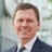 Portrait of Dan Ayley, Global Head of Industrial, Science and Technology, standing outside in front of an outdoor urban setting, featuring modern architectural elements with clean lines. There are hints of greenery, suggesting some trees or plants.