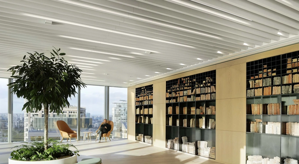 Bright plant filled office space with glass windows and shelf full of books. There's a man in the corner on a orange chair.