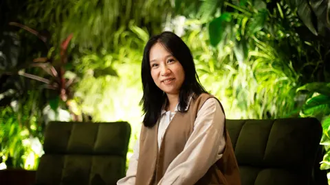 Portrait of Gillian Wang, Graduate Cost Manager, Australia, sitting on dark green chair surrounded by trees and bushes.