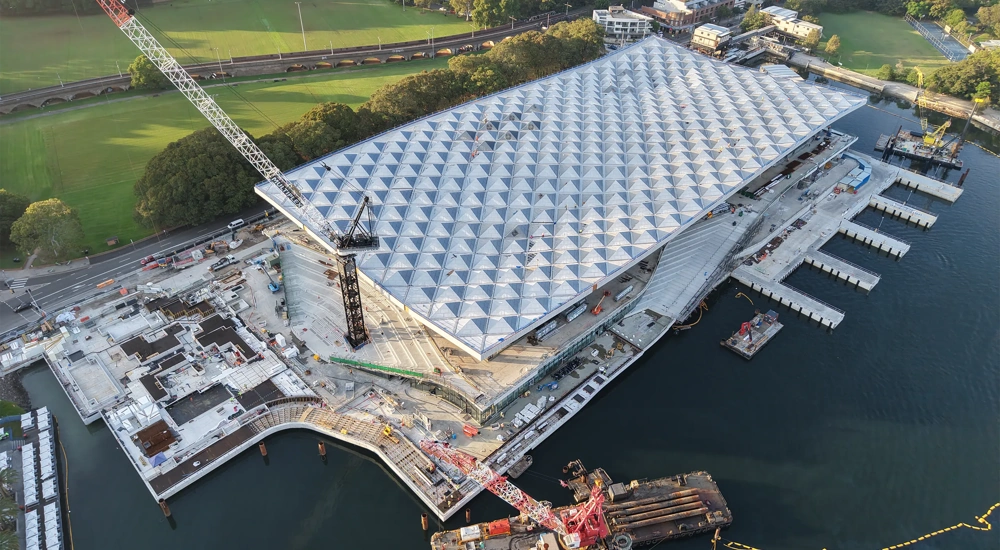 Ariel view of a large waterfront construction site with a geometric roof structure and surrounding cranes and platforms