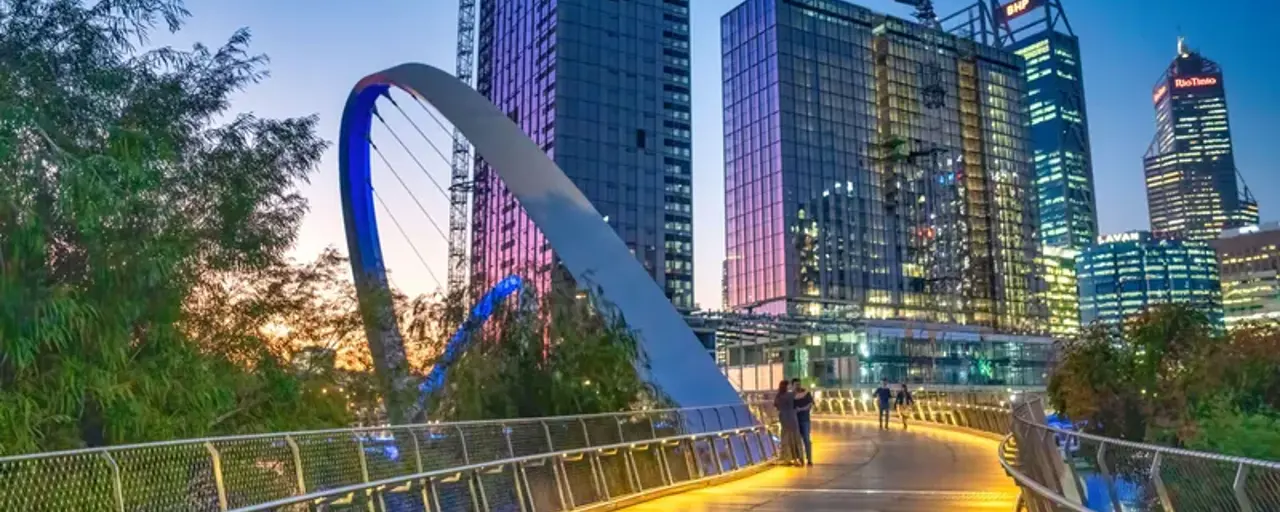 A scenic evening view of a modern pedestrian bridge with curved architecture, illuminated by warm yellow lights, leading towards tall glass skyscrapers reflecting the twilight sky. Lush greenery surrounds the walkway, while the skyline features cranes and iconic buildings