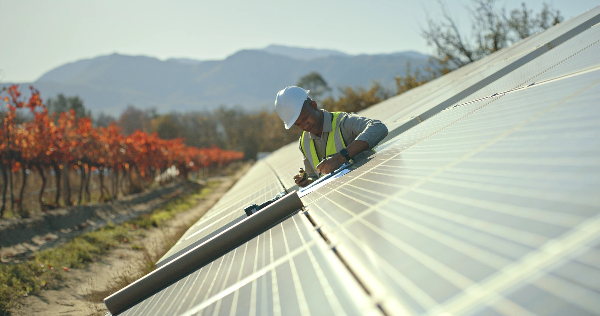 A workman working on a solar panel installation with a backdrop of a mountain and trees with orange and red leaves.