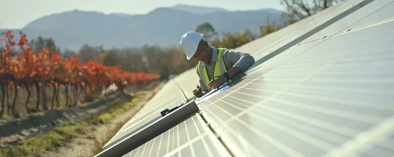 A workman working on a solar panel installation with a backdrop of a mountain and trees with orange and red leaves.  