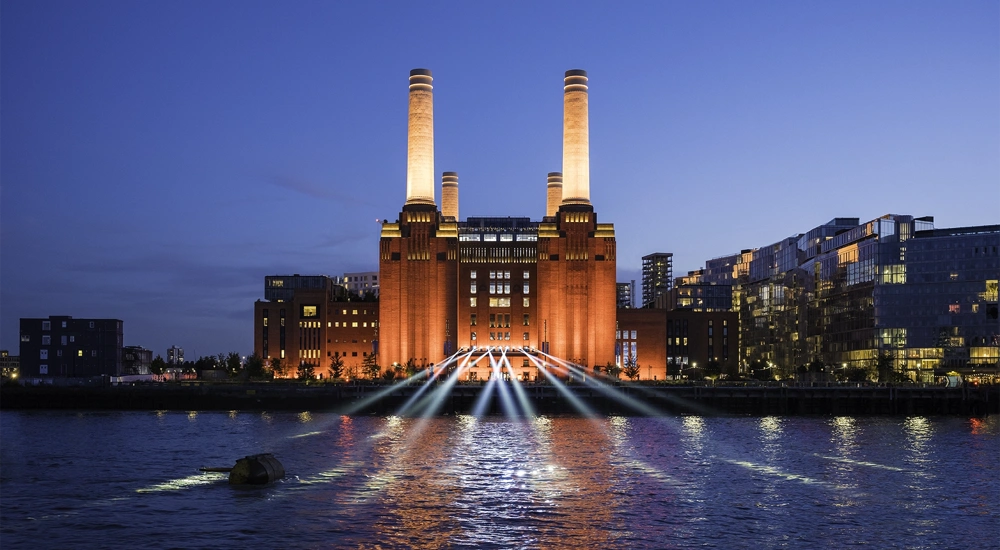 A striking image of London's iconic Battersea Power Station against a blue night sky. The industrial columns are lit up and light also shines on the water in front. To the right of the building are a block of modern, glass fronted flats.