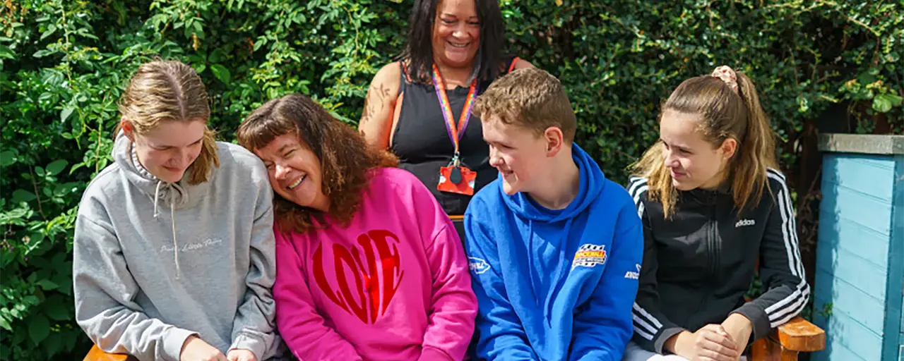 Picture of four children sitting on a bench outside in pink, blue, black and grey clothing, with a woman standing behind the bench.