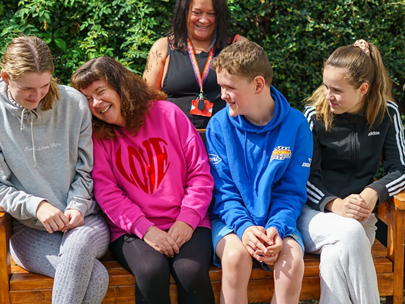 Picture of four children sitting on a bench outside in pink, blue, black and grey clothing, with a woman standing behind the bench.