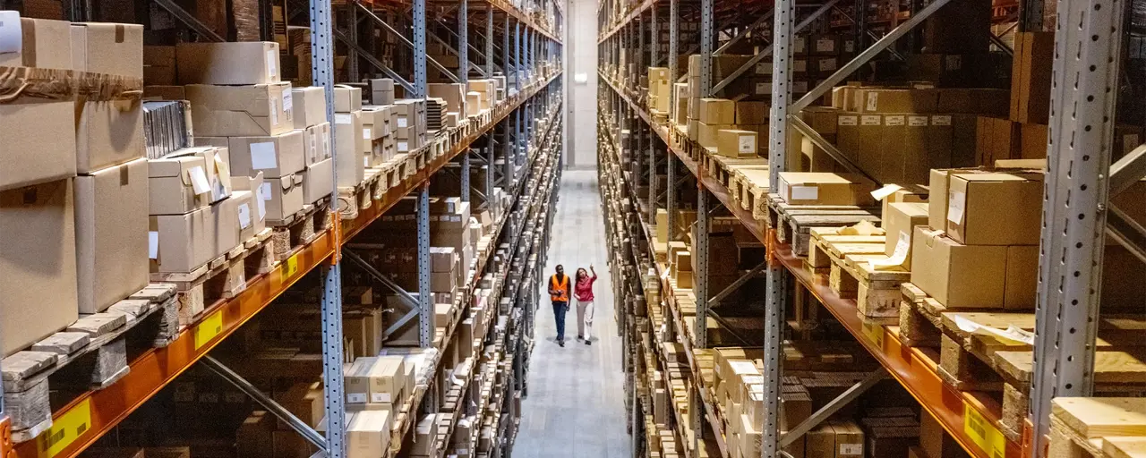 Two employees in high vis jackets walking through warehouse with tall shelves full of products