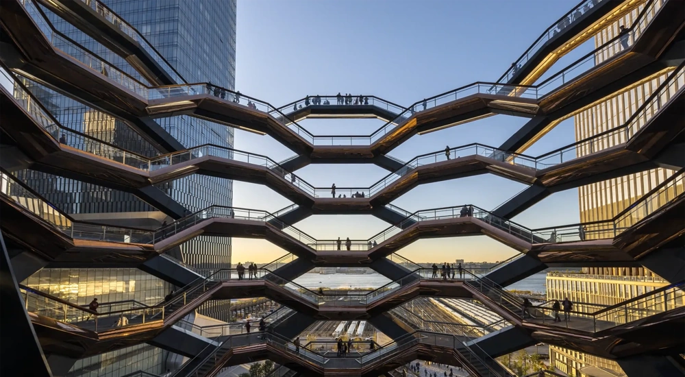 Architectural view of the Vessel at Hudson Yards in New York City, showing multiple levels of interconnected staircases with silhouetted people walking against a backdrop of modern skyscrapers and a sunset sky