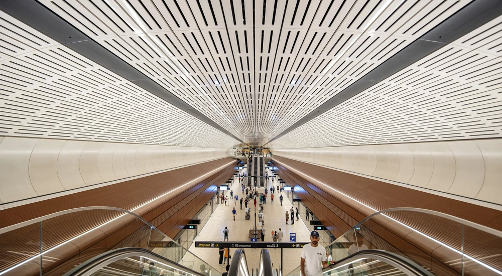 Interior view of Sydney metro modern underground station with a wide ceiling featuring linear ventilation panels, symmetrical architecture and passengers moving through space below.