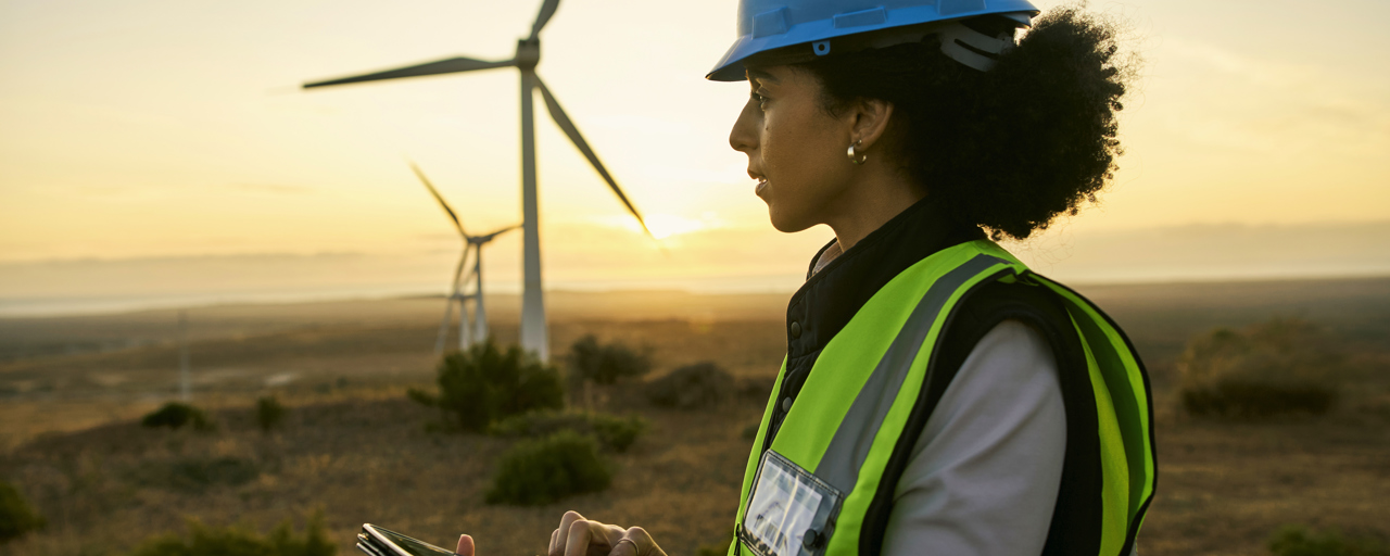 A female engineer in a blue hard hat and high-visibility vest is standing in front of wind turbines at sunset, using a tablet for data collection or monitoring. She is focused on her work, with the wind turbines symbolizing renewable energy and sustainable technology in the background.