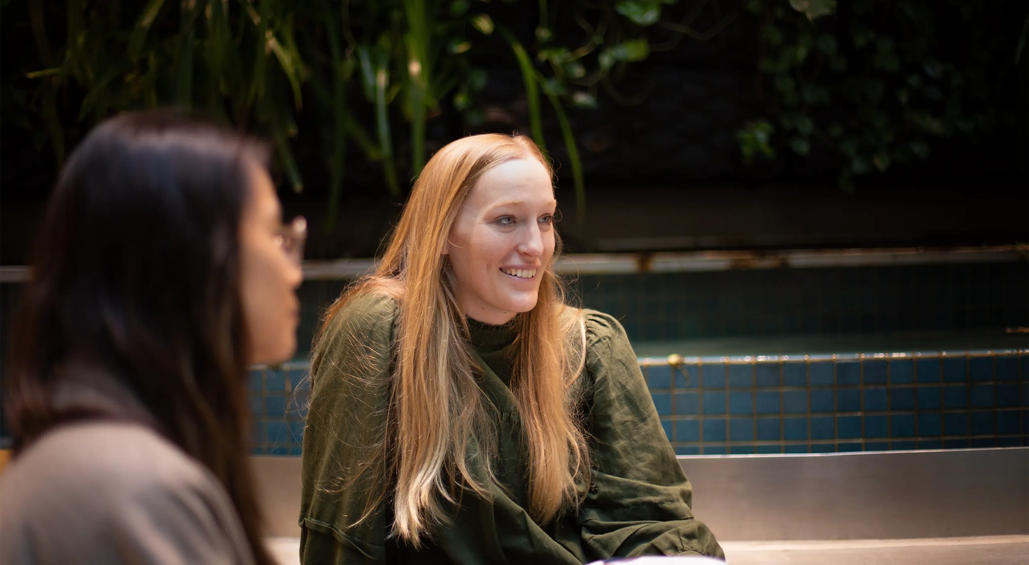 Two colleagues together with the main focus featuring a blonde woman having a conversation in a plant-filled office
