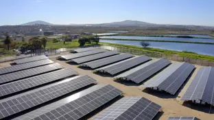 A collection of solar panels next to a body of water with mountains in the background. 