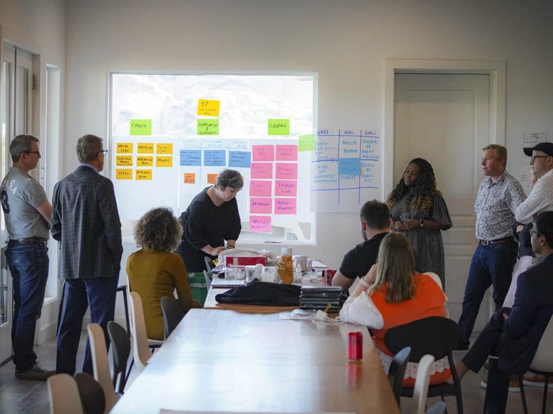 Photo of a group of people seated and standing around a table in a boardroom during a meeting, with a project screen behind them displaying different coloured blocks of text in the form of an organisational chart