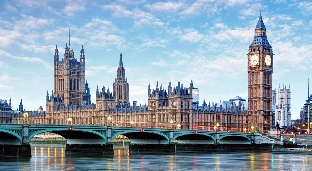 Houses of Parliament, in London, England. Taken from across the River Thames, capturing the entirety of the historic building, including the Elizabeth Tower, which houses Big Ben, on the right. The Westminster Bridge spans the river in the foreground, with the calm water reflecting the structure above. The sky is partly cloudy, and the scene is bathed in soft daylight.