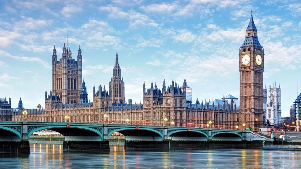 Houses of Parliament, in London, England. Taken from across the River Thames, capturing the entirety of the historic building, including the Elizabeth Tower, which houses Big Ben, on the right. The Westminster Bridge spans the river in the foreground, with the calm water reflecting the structure above. The sky is partly cloudy, and the scene is bathed in soft daylight.