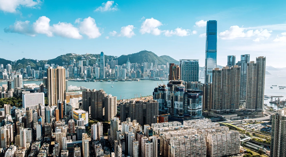 Urban skyline of Hong Kong city featuring high-rise buildings and the harbor surrounded by mountains and blue skies.