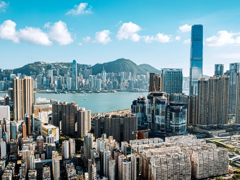 Urban skyline of Hong Kong city featuring high-rise buildings and the harbor surrounded by mountains and blue skies.