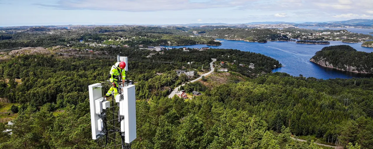 Mobile 5G installation, with man in hi vis on top of 5G tower, surrounded by trees
