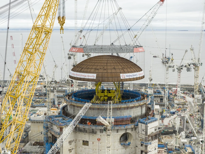 A large crane lifts a domed steel structure into place above a major construction site with multiple cranes and industrial buildings.
