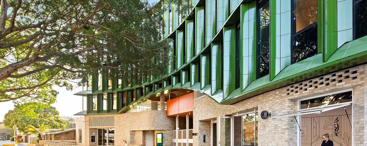 The exterior of a modern, green building with foliage and seating next to it. 