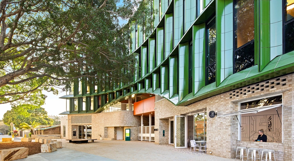 The exterior of a modern, green building with foliage and seating next to it.