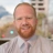 Portrait of Chris Robinson, Associate Director, Contract Services, outside in front of a blurred brown background featuring a building and mix of natural and urban elements.
