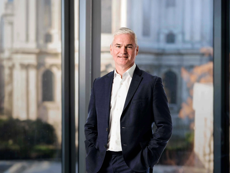 Vincent Clancy standing in the London office with the London city view in the background