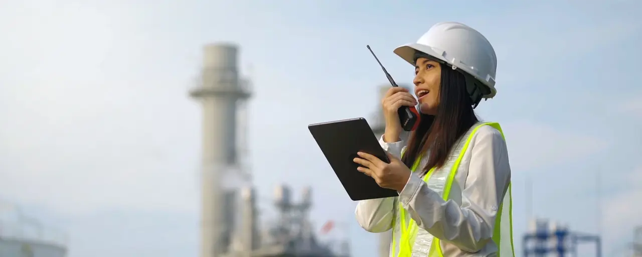 Woman with hard hat in high vis vest talking on a walkie talking at a plant
