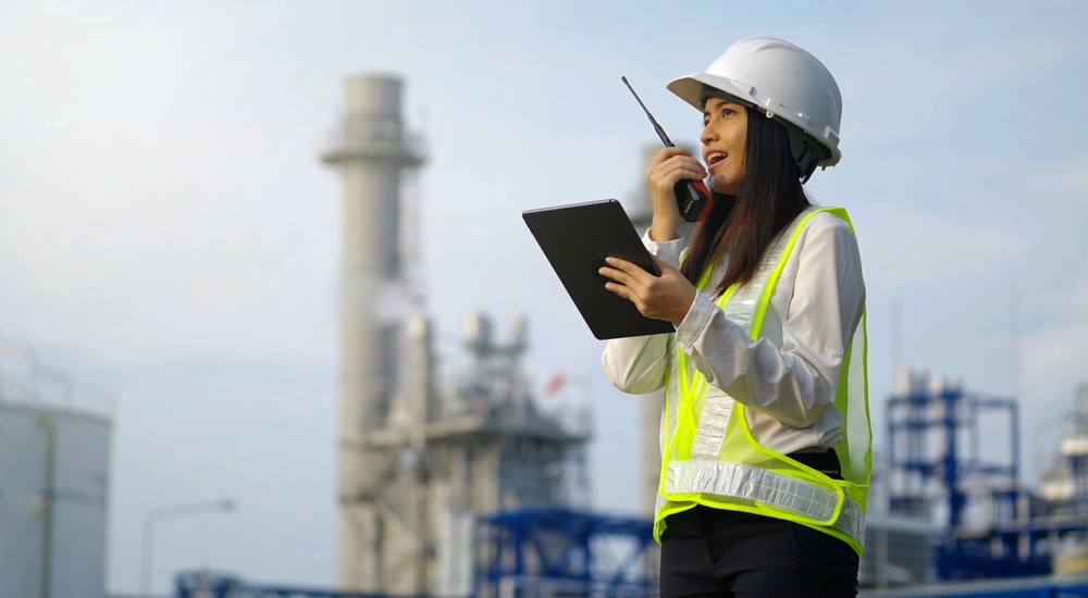 Woman with hard hat in high vis vest talking on a walkie talking at a plant