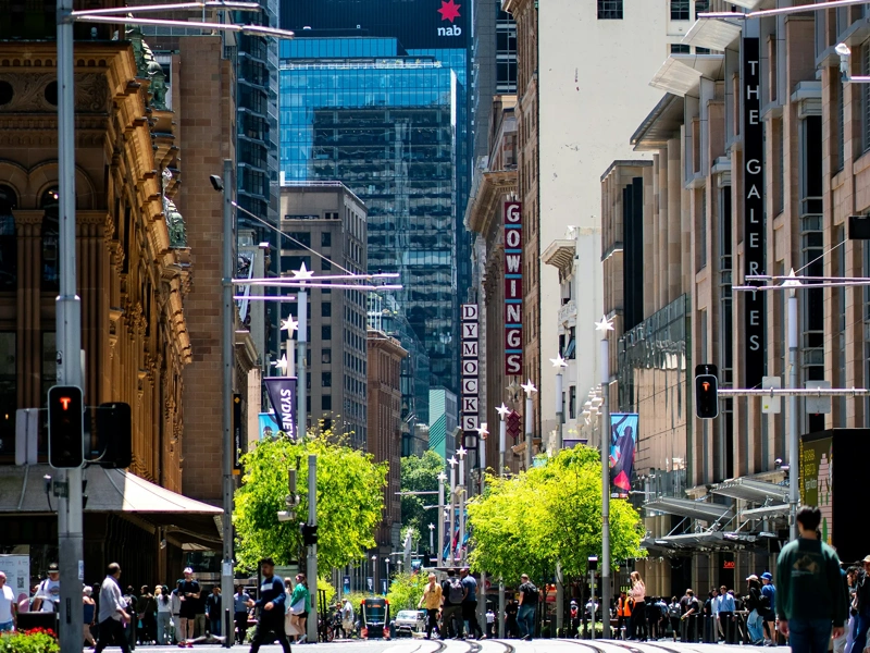 City landscape with people walking and trees in the center with tram coming down in distance.